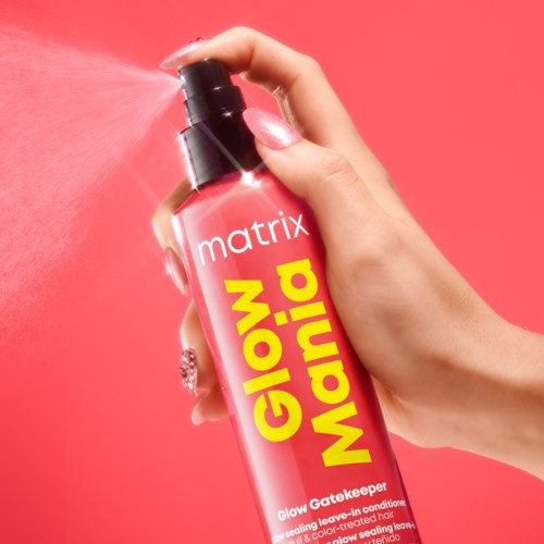 En hånd sprayer rosa hårspray med en gul og rosa etikett for merket Matrix på en rosa bakgrunn.