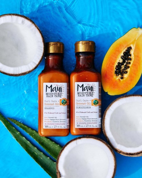 Twee kleine flessen Maui haarverzorgingsproducten omgeven door halve kokosnoten, papaya en aloë vera op een blauwe achtergrond.
