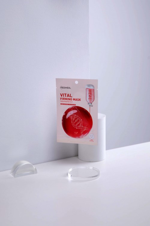Een huidverzorgingsproduct genaamd 'Vital Firming Mask' in rood en wit verpakking, geplaatst op een witte oppervlakte met kleine standaard.