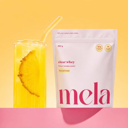 En rosa og gul bakgrunn med et glass med ananas og sitroninfusjon ved siden av en hvit pose med proteinpulver.