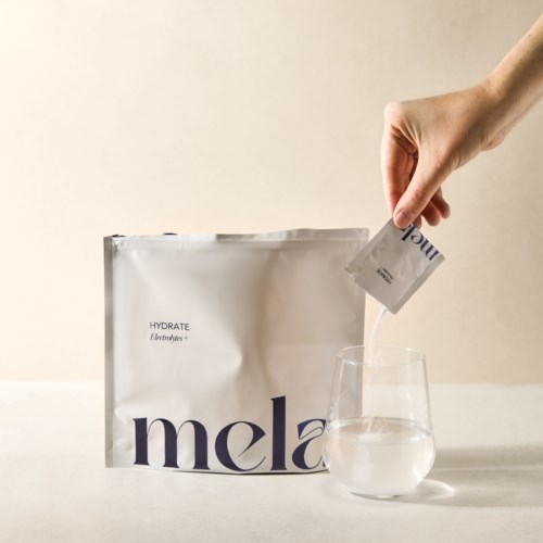 A person pouring powder from a sachet into a glass of water. There is a white bag with purple text next to it.