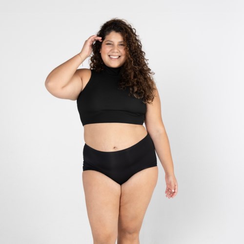 Eine lächelnde Frau mit lockigem Haar trägt ein schwarzes Crop-Top und hoch sitzende Unterwäsche, steht vor einfachem Hintergrund.