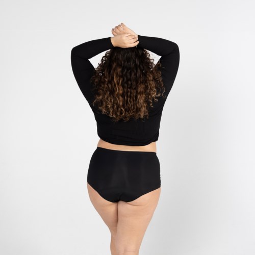 Eine Frau mit lockigem Haar steht mit dem Rücken zur Kamera, Hände auf dem Kopf, trägt schwarze Kleidung.