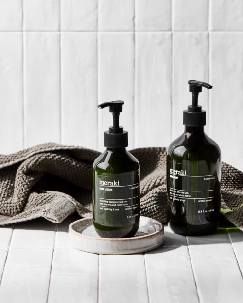 Two dark green bottles with pumps, labeled 'meraki', on a white dish in front of a towel and white tiles.