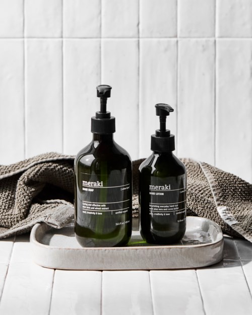 Two dark green bottles with pumps labeled 'meraki' are on a white tray. There is a towel and white tiles in the background.