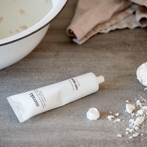 Valkoinen putkilopullo, jossa lukee 'Foot Cream', on puupöydällä, ja vieressä on ripoteltu jauhe ja moppimattos.