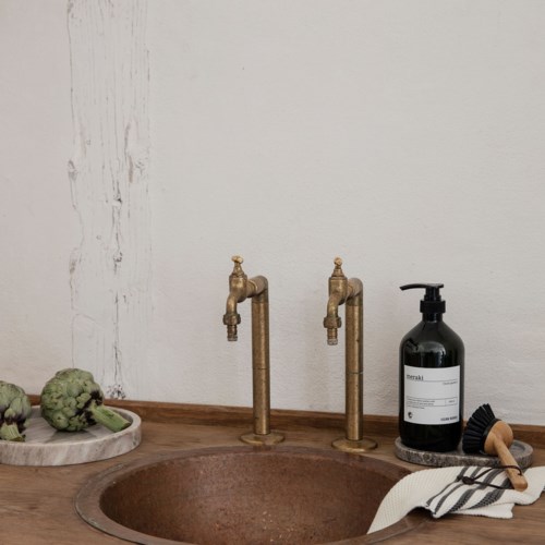 Two vintage-style brass faucets, a black soap dispenser, and some artichokes on a wooden tray on a sink.