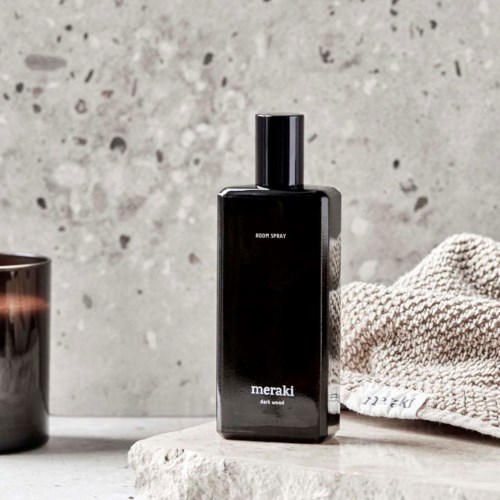 Eine schwarze Flasche mit der Aufschrift 'Meraki room spray' auf einer grauen Oberfläche, neben einem Handtuch und einem kleinen Behälter, vor einer Betonwand.