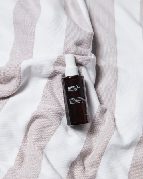 A dark bottle with a spray top on a white and beige striped fabric. The bottle has a label with the word 'meraki.'