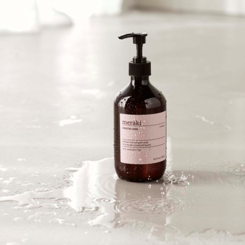 A brown bottle with a pump, labeled 'Meraki', sits on a wet surface with water splashes around it.