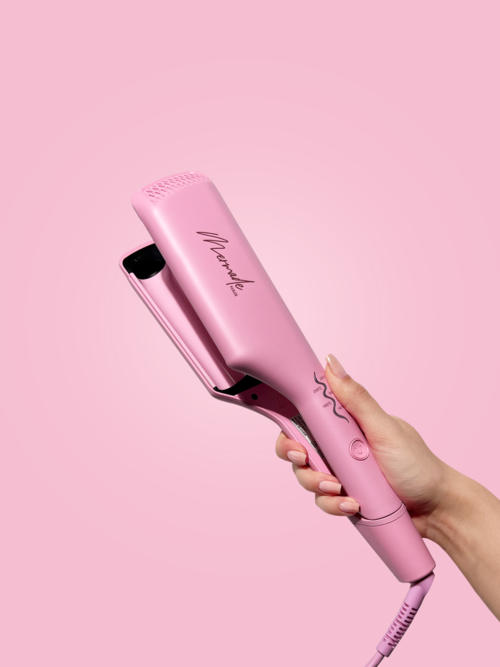 A hand holds a pink hair straightener against a pink background.