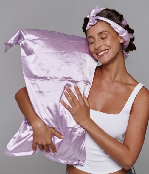A smiling woman in a white tank top holds a large purple satin pillow, wearing a satin ribbon headband.