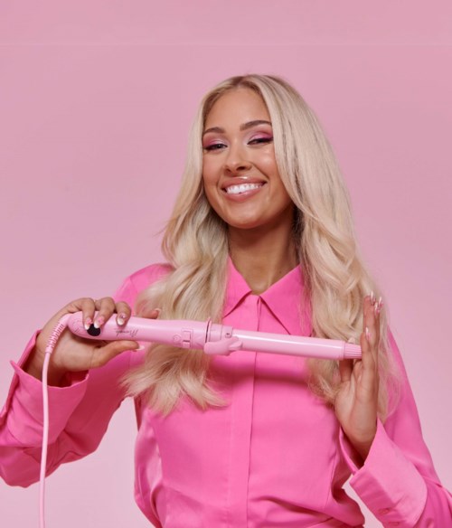 A woman with long blonde hair in pink clothes curls her hair with a pink curling iron.