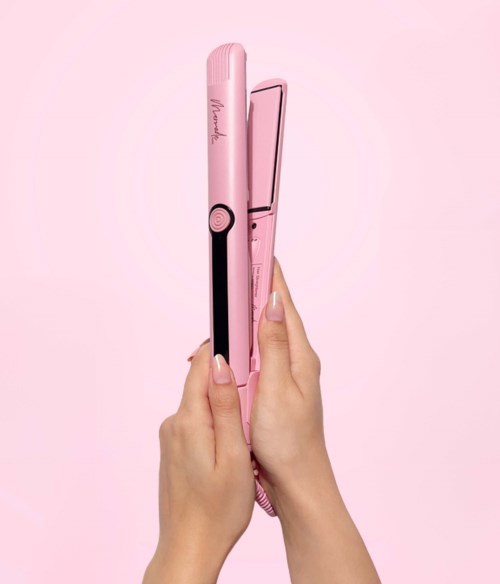 A person holds a pink hair straightener against a pink background.