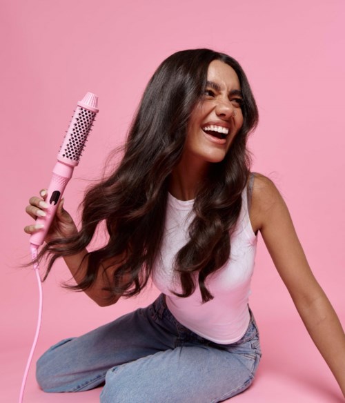 A woman with long dark hair laughing, holding a pink curling iron, against a pink background.