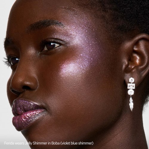 Een vrouw met donkere huid draagt glanzende paarse lippenstift, sprankelende paarse oogschaduw en oorbellen. Haar is kort en gestyled.