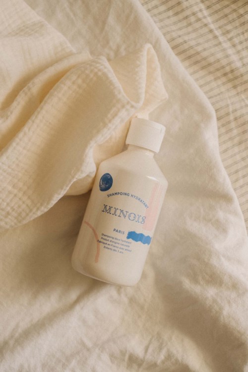 A white bottle of shampoo lies on a white bed with crumpled bedsheets.