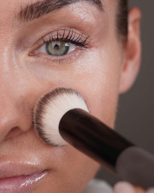 Nærbilde av en kvinne som påfører makeup med børste på kinnet, viser klar hud og detaljert øyemakeup.