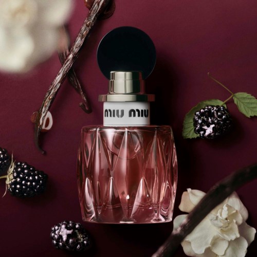 En Miu Miu parfymeflaske omgitt av bjørnebær, hvite blomster og grener på mørk burgunder bakgrunn.