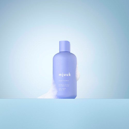 Een paarse fles met huidverzorgingsproduct genaamd 'mjuuk' staat op een oppervlak met schuim ernaast, tegen een zachte blauwe achtergrond.
