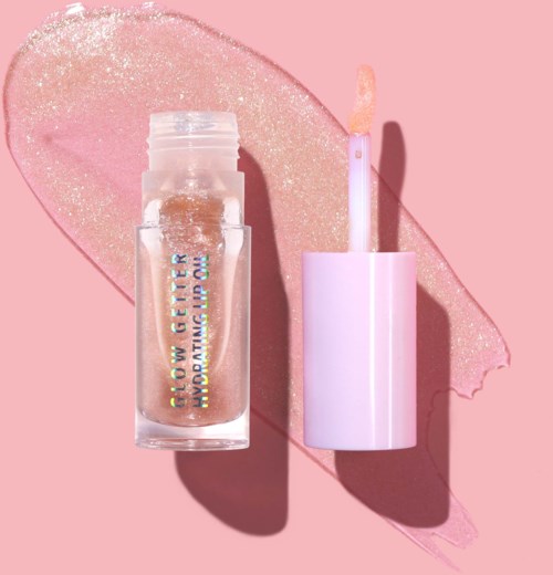 A lip gloss tube with shiny gold gloss inside, placed next to a pale pink lip gloss applicator on a pink background.