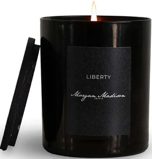 A large black candle with a flame, label says 'Liberty' and 'Morgan Madison,' with a wooden lid beside it.