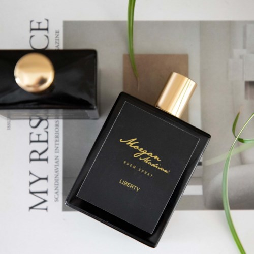 A black perfume bottle with gold accents and modern text, placed on a magazine with plants nearby.