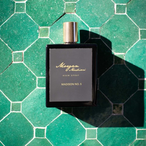 A black perfume bottle with a gold cap lies on green tiled floor, casting a shadow.