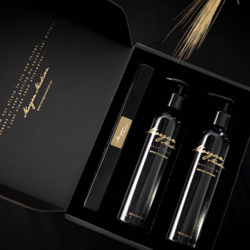 Two transparent bottles with pumps, a black brush, and gold lettering in a black box. Gold sprinkles fall from above.