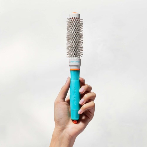 A hand holds a round hairbrush with a teal handle and white bristles. The background is plain and light-colored.