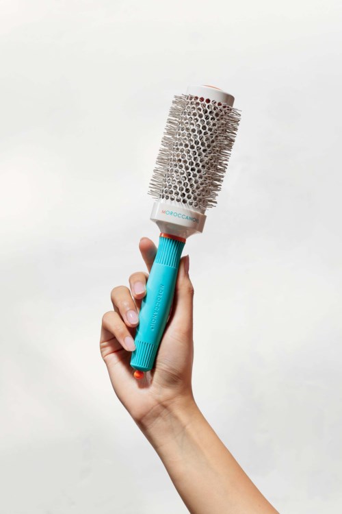 A hand holds a round hairbrush with a blue handle and a white and gray bristle section.
