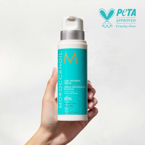 A hand holds a blue and white bottle of Moroccanoil Curl Defining Cream, labeled as cruelty-free and PETA approved.