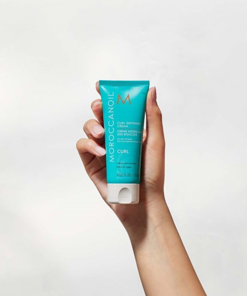 Käsissä on sininen putki Moroccanoil Curl Defining Creamia valkoista taustaa vasten.