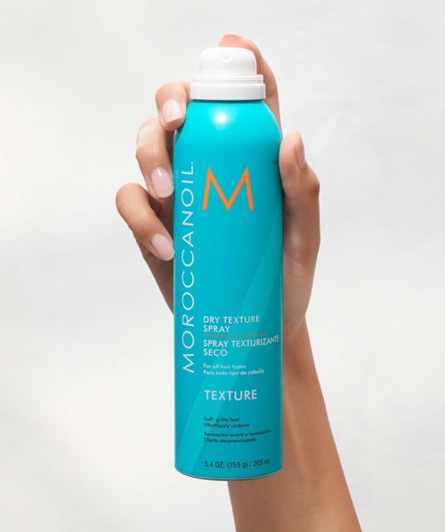 A hand holding a blue can of Moroccanoil dry texture spray against a white background.