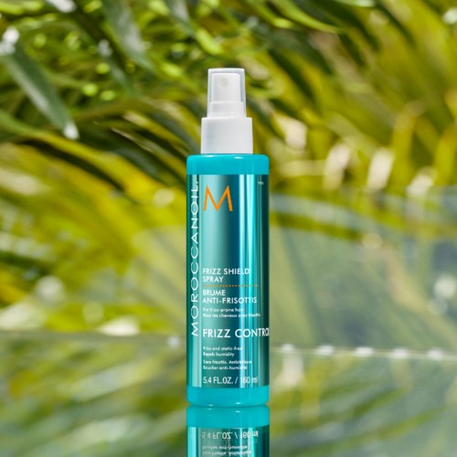 A blue spray bottle labeled Moroccanoil Frizz Control with a spray nozzle, set on a reflective surface with green leaves behind.