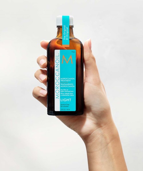 Hand holding brown bottle with blue label for MoroccanOil treatment, Light. Simple background.
