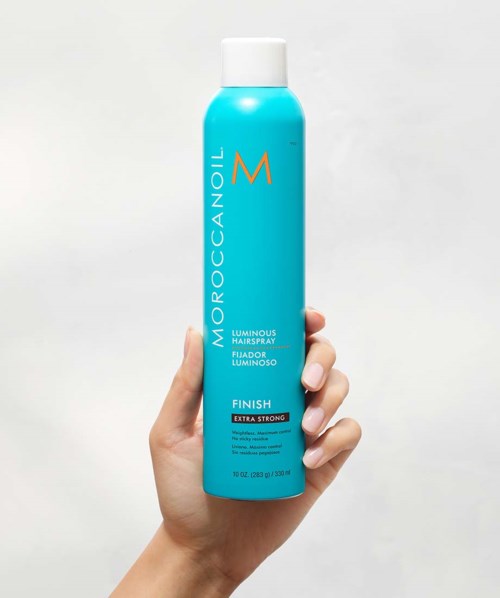 A hand holds a tall turquoise bottle of Moroccanoil hair product with white cap, label reads 'luminous hair spray' and 'extra strong.'