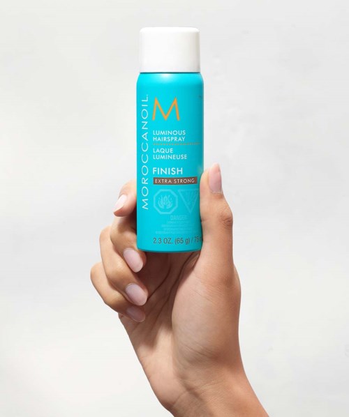 A hand holds a blue bottle of Moroccanoil Luminous Hairspray against a white background.
