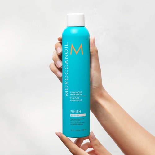 A hand holds a tall, blue can of MoroccanOil Luminous Hairspray with a white cap.