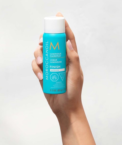 Een hand houdt een blauwe fles Moroccanoil Luminous Hairspray met witte dop.