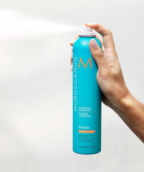 A hand sprays hair product from a blue can, labeled Moroccanoil, on a plain background.