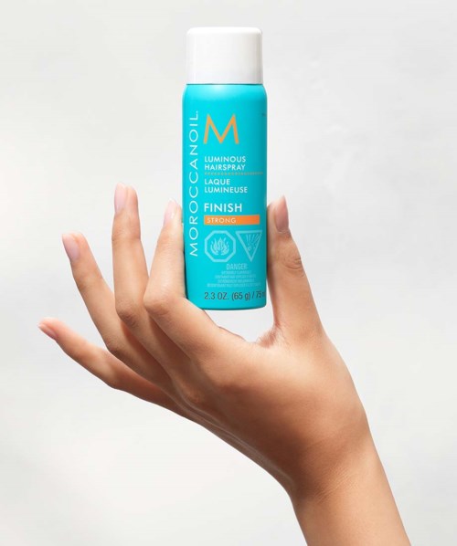 Käsi pitää sinistä Mochaccino Luminous Hairspray -pullot, jossa on valkoinen korkki, taustalla yksinkertainen.
