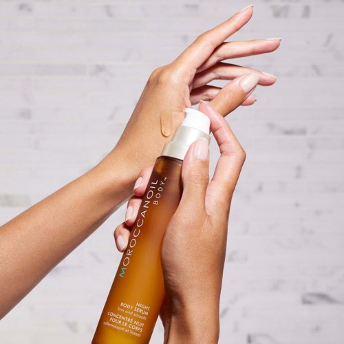 Käsi suihkuttaa ruskeaa pulloa Moroccanoil Body Night Serumia, jossa on valkoinen suihkukaula ja teksti pulloissa.
