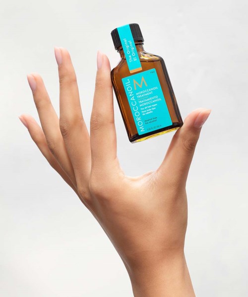 A hand holds a small brown bottle with a blue label and black cap, labeled "Moroccanoil".