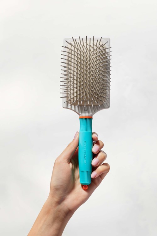 A person holds a blue hairbrush with a rectangular plastic base and metal bristles.