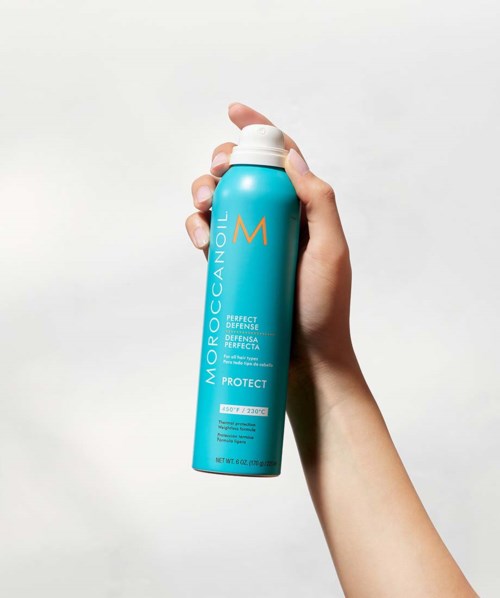 A hand holds a blue bottle of Moroccanoil Protect spray. The bottle has white and orange text and a pump top.