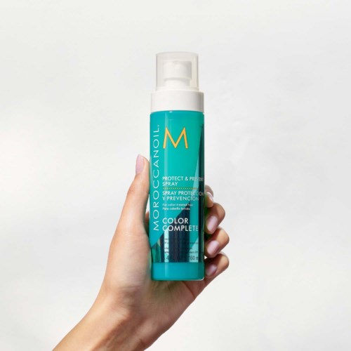 A hand holds a blue and white spray bottle labeled Moroccanoil Color Complete Protect & Prevent.