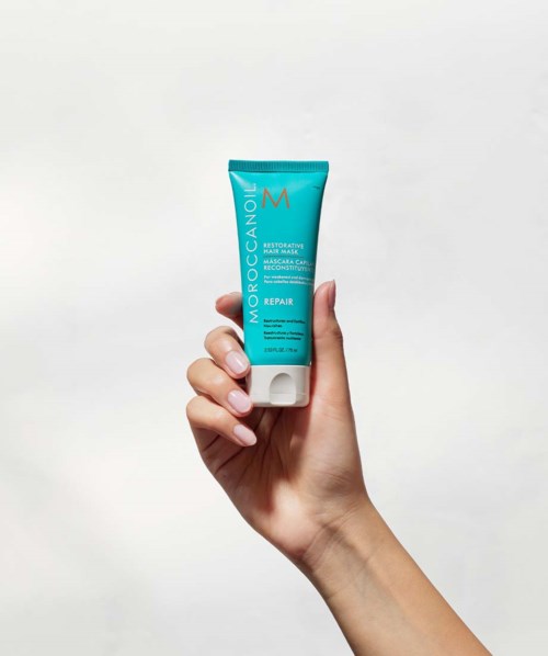 Käsi pitää sinistä Moroccanoil-hiustuoteputkea valkoista taustaa vasten.