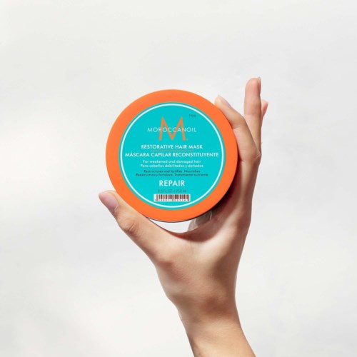 A hand holds a round container with a blue label, orange border, and white text for hair mask product.