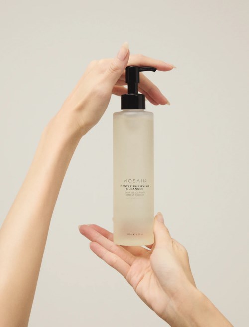 Två händer håller en beige flaska med svart pump, märkt MOSAIC Gentle Purifying Cleanser.